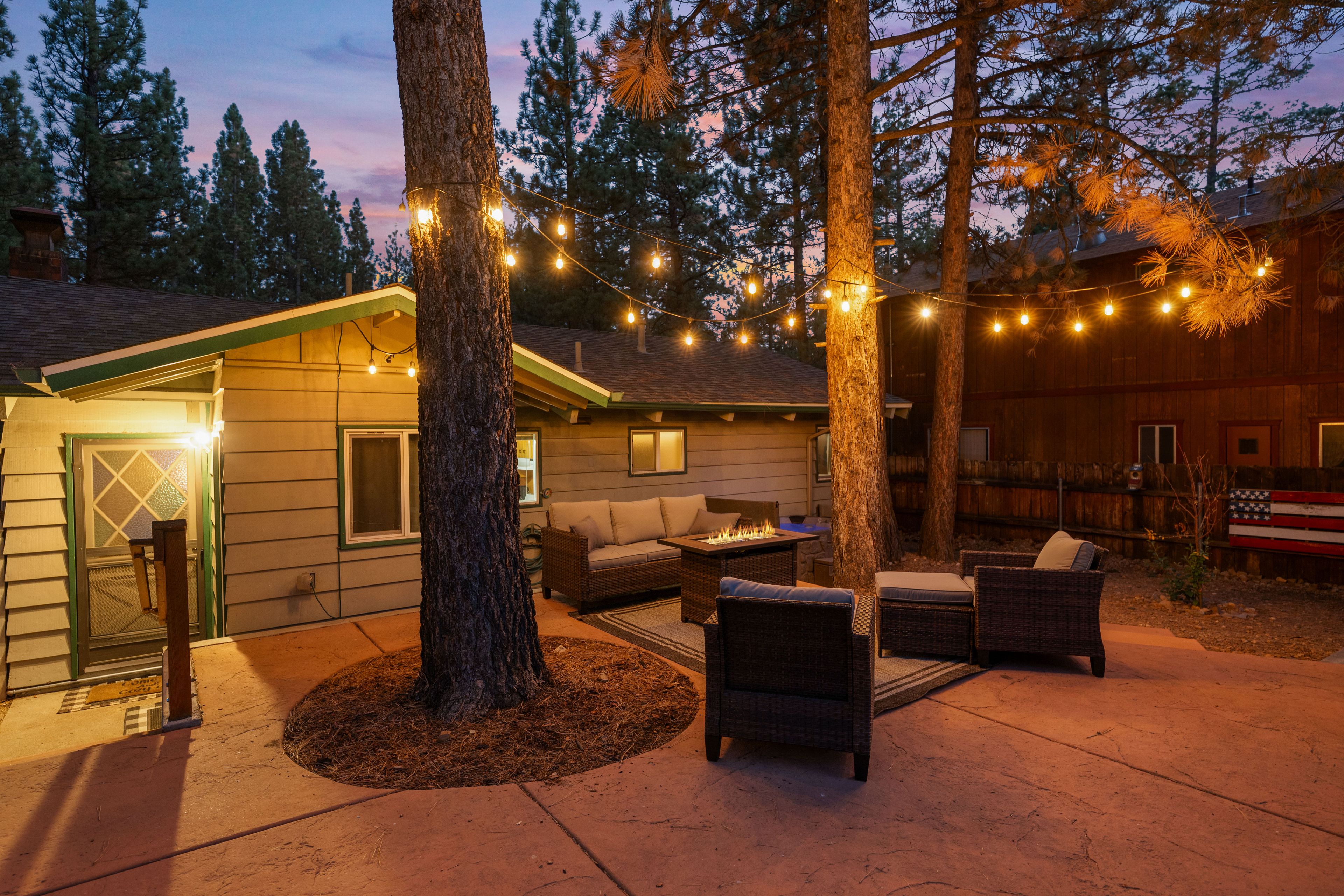 Un espacio al aire libre acogedor con una chimenea, rodeado de árboles y luces colgantes.