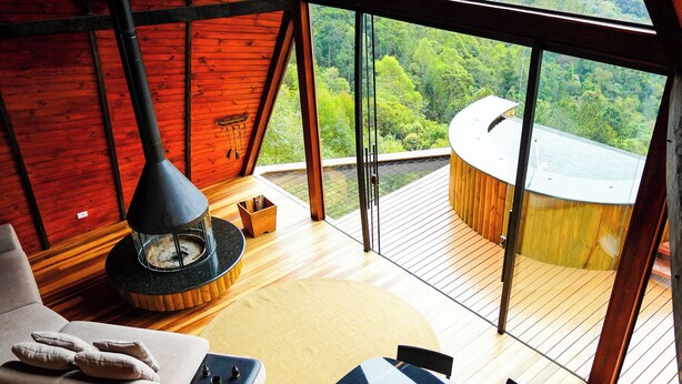 Een gezellig chaletinterieur met een houten vloer, een eettafel en een bank. Een groot raam biedt uitzicht op een bebost landschap.