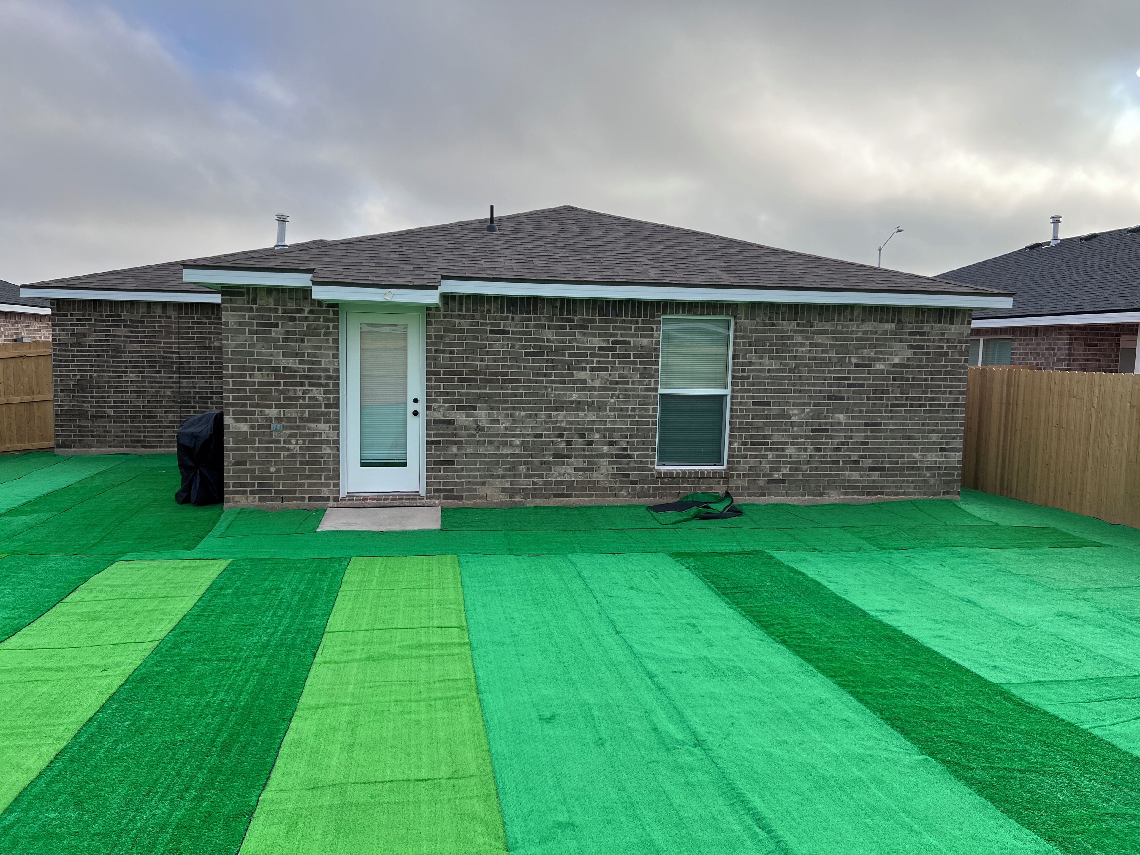 Una casa residencial con fachada de ladrillo, una puerta principal, un ventanal pequeño y un césped artificial verde.