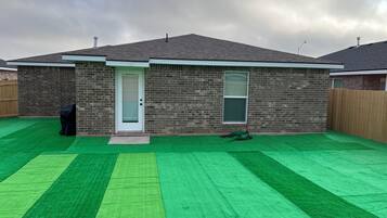 Una casa residencial con fachada de ladrillo, una puerta principal, un ventanal pequeño y un césped artificial verde.