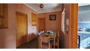 Un comedor con una mesa y sillas de madera, una lámpara de techo y una cocina visible a través de una puerta abierta.