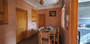 A dining area with a wooden table and chairs, a ceiling light, and a kitchen visible through an open door.