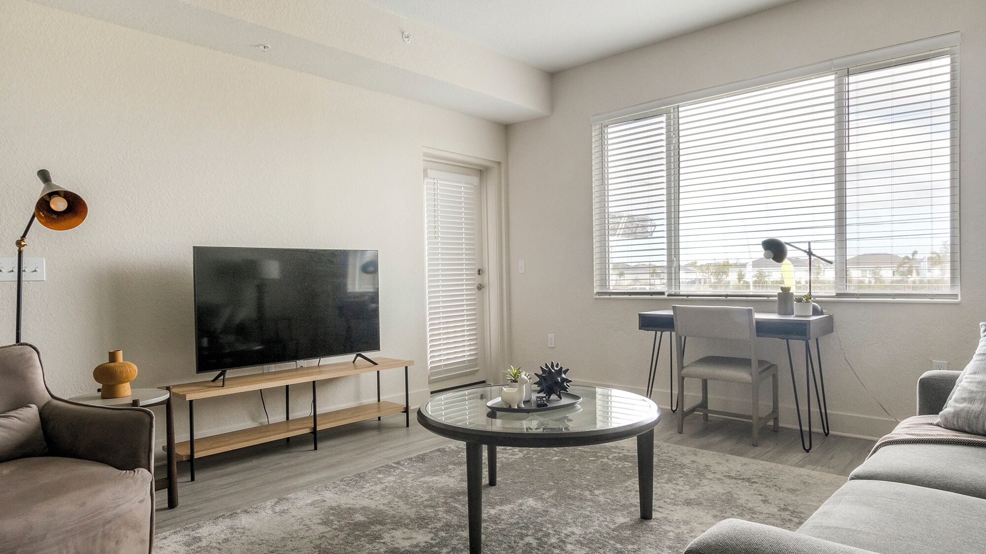 Un salon avec un téléviseur à écran plat posé sur un meuble en bois, une table basse en verre, un canapé gris, un bureau avec une chaise et une grande fenêtre équipée de stores.