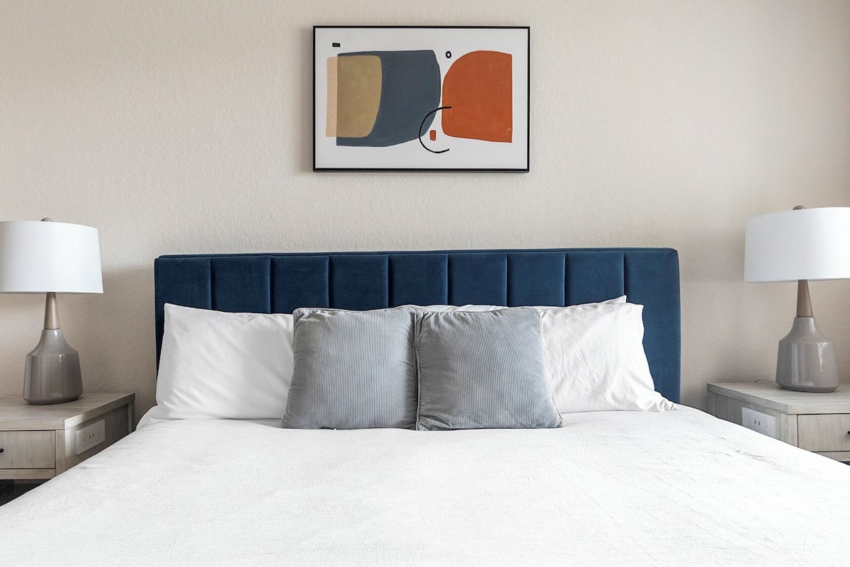 A neatly made bed with a blue upholstered headboard, two bedside tables with lamps, and a framed abstract artwork on the wall.