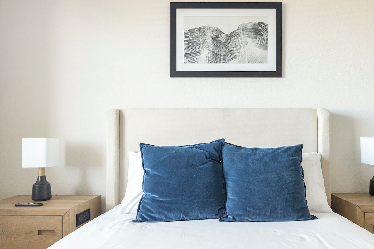 A neatly made bed with two pillows, a framed artwork above it, and two bedside tables with lamps on either side.