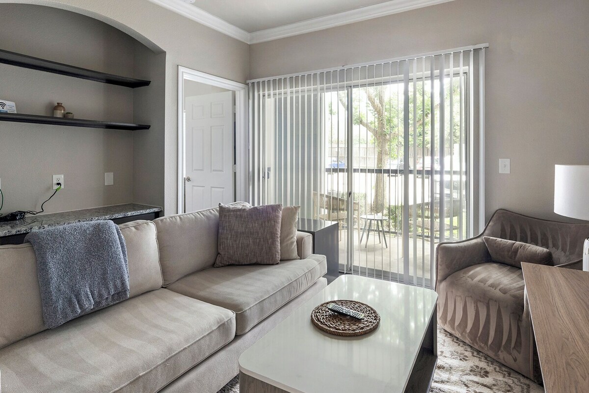 A living room with a sofa, armchair, coffee table, and a lamp. There is a sliding glass door leading to an outdoor area.