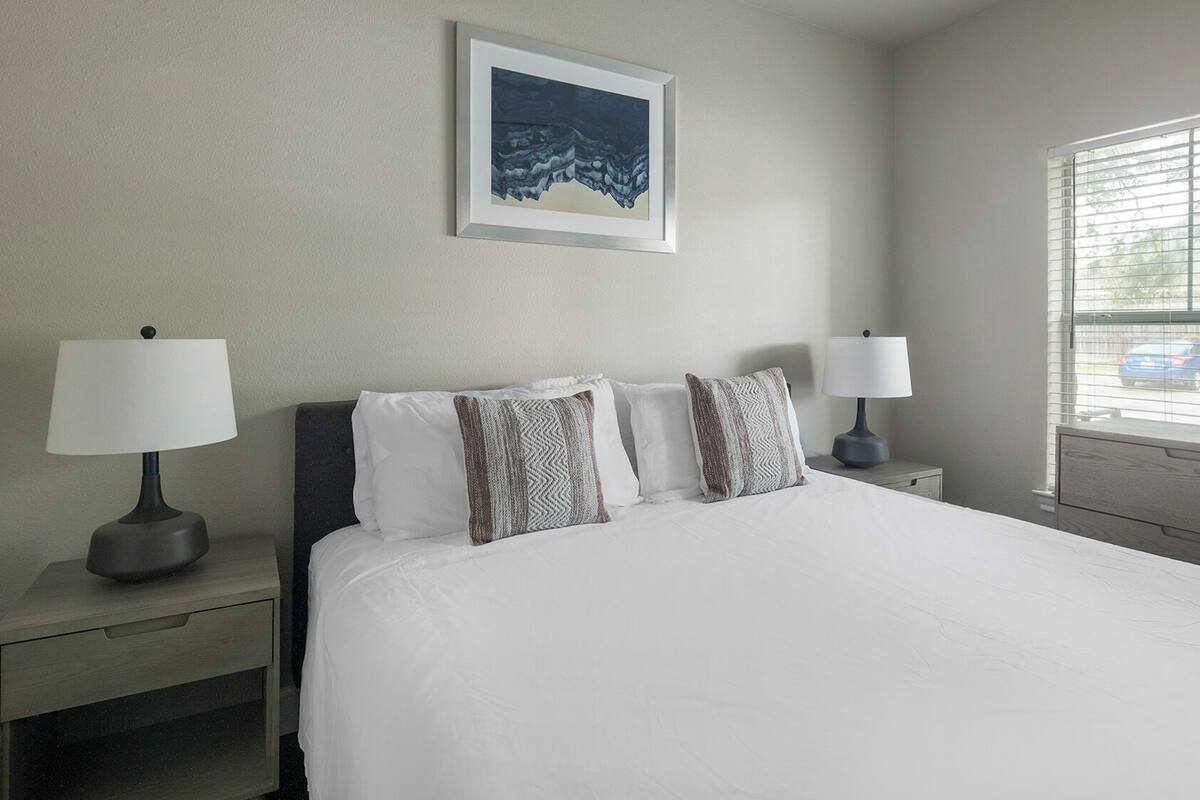 A hotel room with a bed, two bedside tables with lamps, a window with blinds, and a framed picture on the wall.