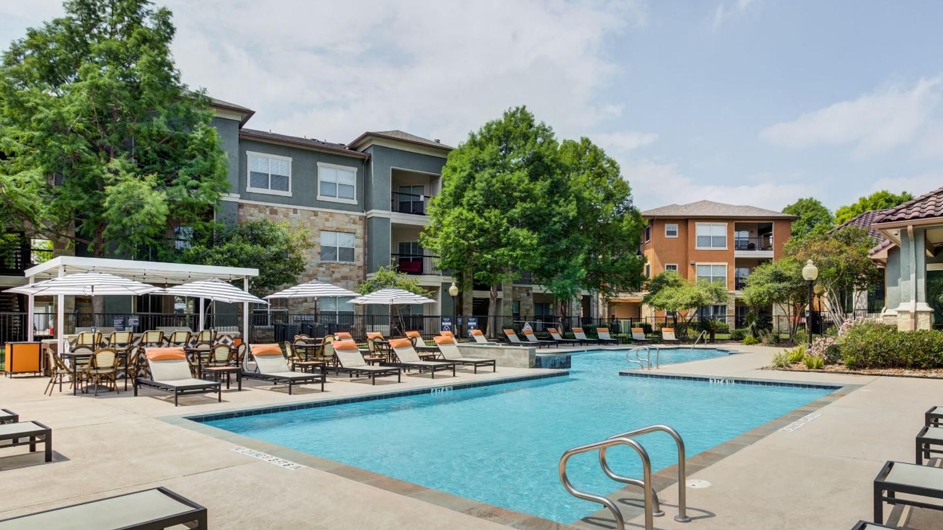 A pool area with lounge chairs and umbrellas, surrounded by multi-story buildings with balconies.