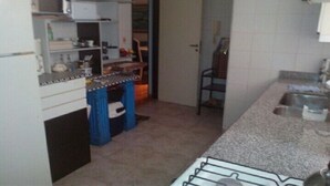 Uma cozinha compacta com armários brancos, micro-ondas e pia. Há uma cadeira e uma cortina azul ao fundo.