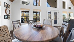 Un coin repas avec une table ronde en bois, des chaises et un saladier rempli de fruits. De nombreuses fenêtres offrent une vue sur l’extérieur.