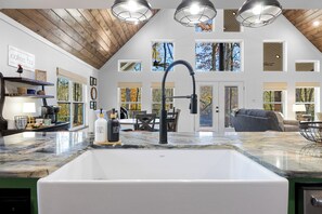 A spacious kitchen with a large white sink, wooden ceiling, and multiple windows offering views of trees.