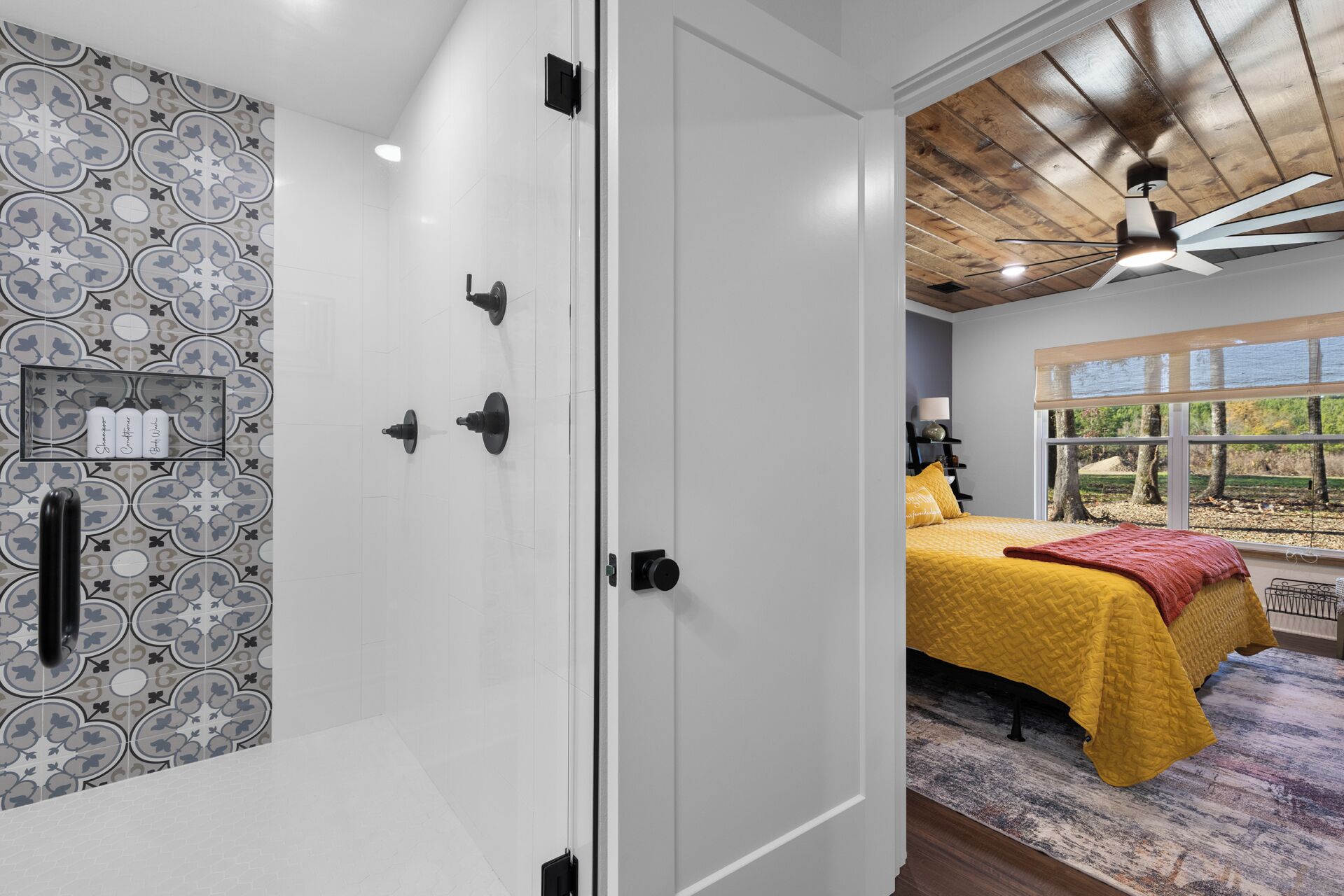 A bedroom with a patterned wall, a bed with a yellow bedspread, a ceiling fan, and a window with a view of trees.