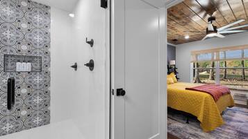 A bedroom with a patterned wall, a bed with a yellow bedspread, a ceiling fan, and a window with a view of trees.