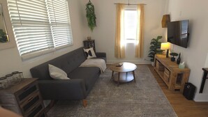 A living room with a grey sofa, a round coffee table, a wooden side table, a TV mounted on the wall, and a window with blinds.