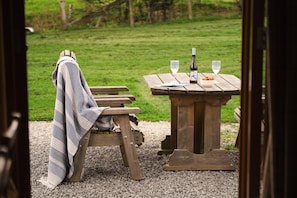 Un banc en bois avec une couverture, une table de pique-nique avec une bouteille et des verres, et un champ verdoyant en arrière-plan.
