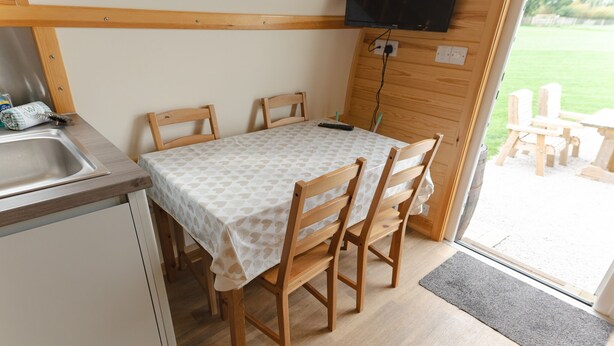 A compact kitchen with a sink, wooden chairs, a table with a patterned tablecloth, and a TV mounted on the wall. - En-suite Wooden Cabin With Hot Tub (Langho)