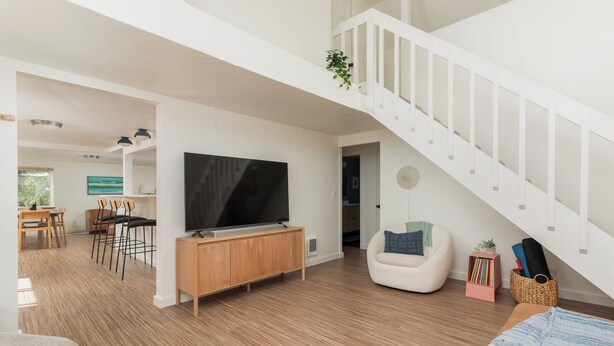 A modern living room with a wooden floor, a large flat-screen TV, a white staircase, and a cozy seating area with a bean bag and a small side table.