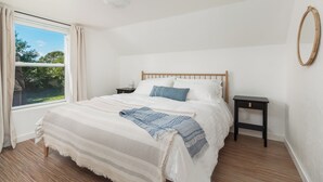 A bedroom with a large bed, a nightstand, a bedside table, a window with curtains, and a mirror on the wall.