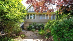 A two-story house with a stone pathway leading to the front door, surrounded by lush greenery and a variety of plants.