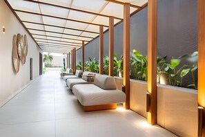 A corridor with a row of sofas and chairs, a glass wall with plants, and a ceiling with a grid pattern. - Way Bossa (João Pessoa)
