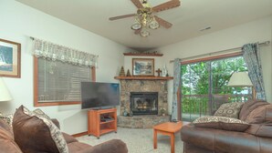 A living room with two brown leather sofas, a stone fireplace, a flat-screen TV, and a ceiling fan.