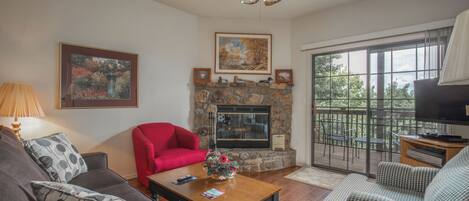 A living room with a stone fireplace, a ceiling fan, and a large window with a view of trees.