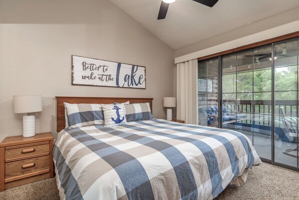 A bedroom with a checkered bedspread, a nightstand with a lamp, a wall-mounted sign, and a view of a balcony with outdoor furniture.