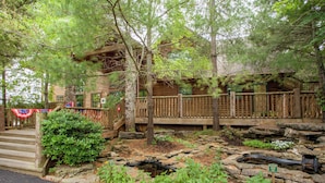 A rustic log cabin with a wooden deck, stone pathway, and a small pond. - 2BR Village At Indian Point 905 near SDC-Free Tickets Daily up to $500 (Branson)
