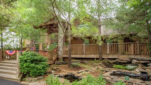 A rustic log cabin with a wooden deck, stone pathway, and a small pond.