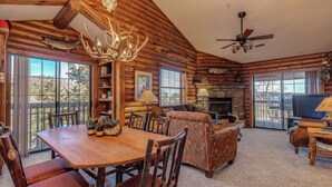 A log cabin interior with a dining area, sofa, TV, fireplace, and large windows. - 2BR Village At Indian Point 1009 near SDC-Free Tickets Daily up to $500 (Branson)