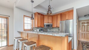 A kitchen with wooden cabinets, a stainless steel refrigerator, a microwave, and a marble countertop. There are three white bar stools.