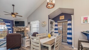 A dining area with a wooden table and chairs, a ceiling fan, a stone fireplace, and a TV mounted on the wall. - 2BR Village At Indian Point 1212 near SDC - Free Tickets Daily up to $500 (Branson)