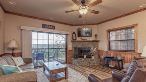 A cozy living room with a fireplace, a ceiling fan, and a large window overlooking a deck. - 2BR Village At Indian Point 1504  near SDC - Free Tickets Daily up to $500 (Branson)