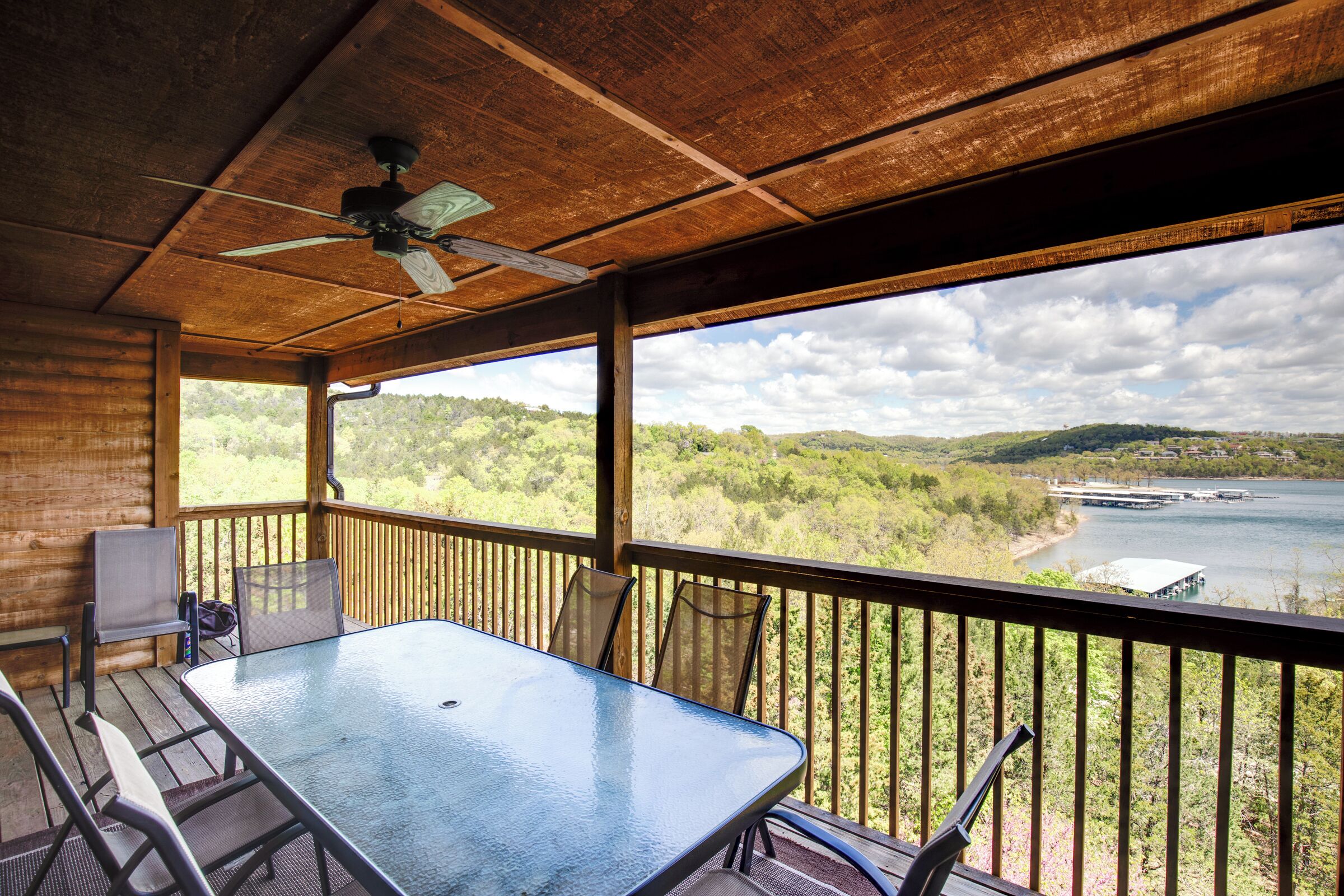 A covered outdoor area with a table and chairs, a ceiling fan, and a view of a lake and forest.