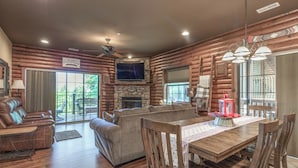 A log cabin interior with a stone fireplace, a television mounted above it, a dining area with wooden chairs and a table, and a ceiling fan. - 2BR Village At Indian Point 2102 near SDC - Free Tickets Daily up to $500 (Branson)