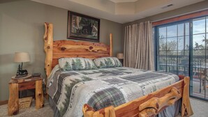 Un dormitorio con cabecera de madera, un ventanal con cortinas, una cama con colcha y una mesita de noche con lámpara.