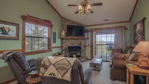 A cozy living room with a stone fireplace, a ceiling fan, and a large window with blinds. - 2BR Village At Indian Point 2009 near SDC - Free Tickets Daily up to $500 (Branson)