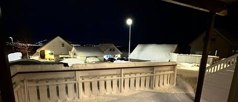 Uma área externa coberta de neve, com cerca de madeira, um poste de luz e edifícios ao fundo.