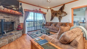 A cozy living room with a stone fireplace, a mounted animal head, a ceiling fan, and a large window. - 2BR Village At Indian Point 1607 near SDC - Free Tickets Daily up to $500 (Branson)