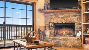 A cozy living room with a stone fireplace, a large window with a view, and a wooden table with a woven mat.