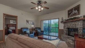 A living room with a stone fireplace, leather chairs, a sofa, a ceiling fan, and a large window with a view of greenery. - 2BR Village At Indian Point 1702 next to SDC- Free Tickets Daily up to $500 (Branson)