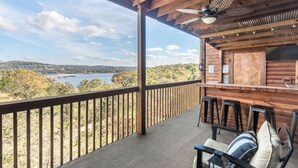 A wooden deck with a view of a lake and hills, a bar area with stools, and a ceiling fan. - 2BR Village At Indian Point 1707 near SDC - Free Tickets Daily up to $500 (Branson)