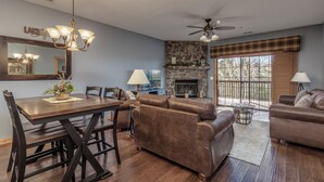 A spacious living room with a fireplace, a dining area with a wooden table and chairs, and a large window with a view of the outdoors. - 2BR Village At Indian Point 1803 near SDC - Free Tickets Daily up to $500 (Branson)