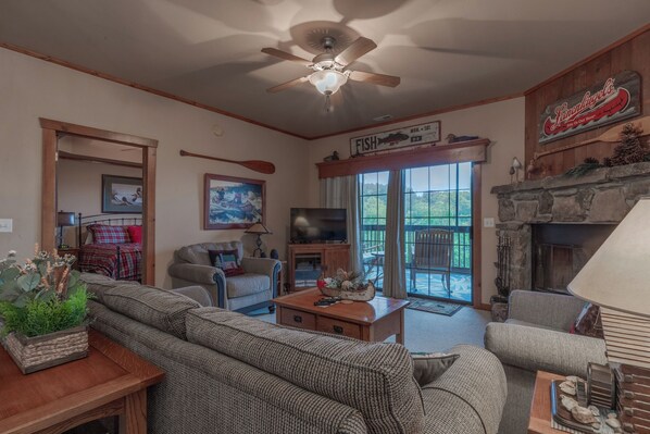 A cozy living room with a fireplace, a ceiling fan, and a large window overlooking a deck.
