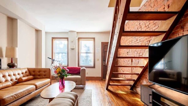 Una sala de estar moderna con un sofá de cuero, una mesa de centro y un televisor. Se observa una escalera con paredes de ladrillo visto y vigas de madera.