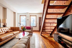 A modern living room with a leather sofa, a coffee table, and a TV. There is a staircase with exposed brick walls and wooden beams.