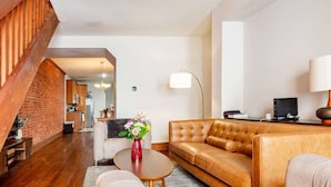 Una sala de estar moderna con un sofá de cuero marrón, un sillón blanco y una mesa de centro con un jarrón de flores. Se ve una pared de ladrillo y una escalera de madera al fondo.