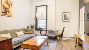 Una sala moderna con sofá, mesa de centro, mesa de comedor y sillas. Hay un cuadro en la pared y una lámpara junto a la ventana.
