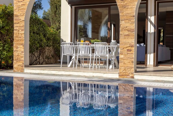 Un espace aménagé au bord de la piscine, avec une table et des chaises, un pilier en pierre et un reflet dans l’eau.