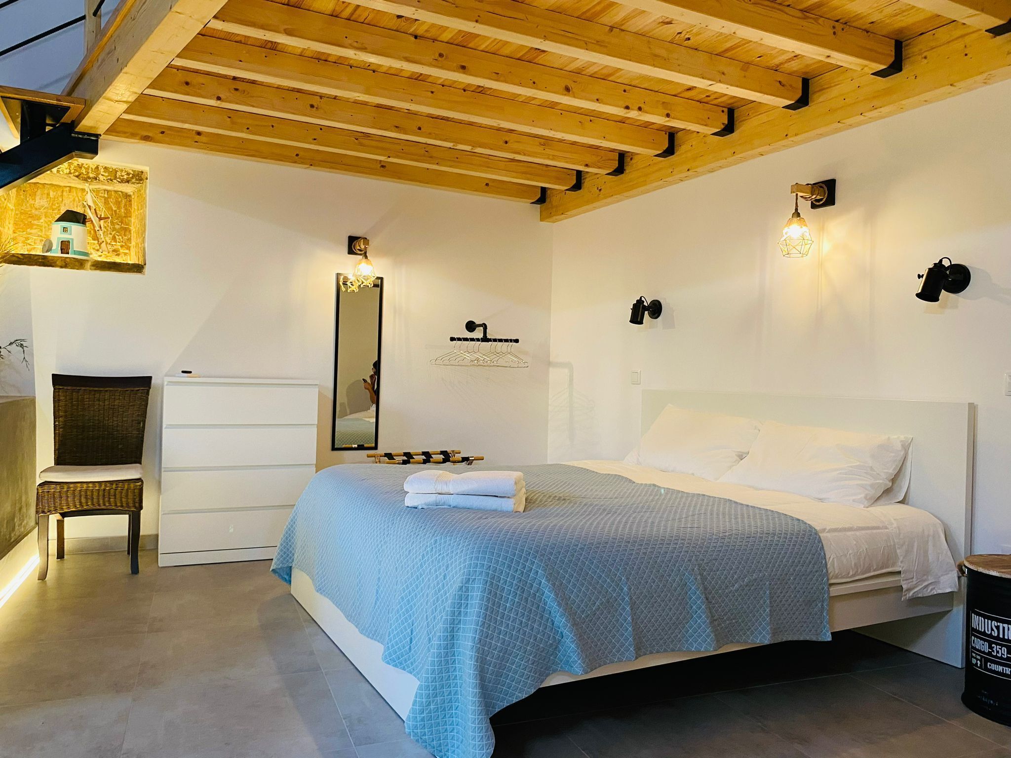 Un dormitorio con cama, una silla, cómoda, espejo e iluminación en la pared.